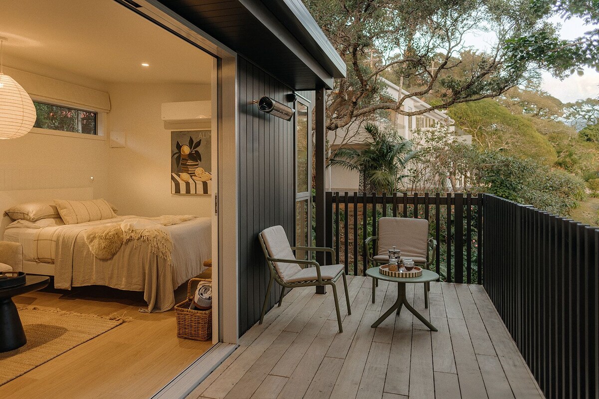 A bedroom with a bed, bedside table, and a window. There is a balcony with a chair and a table.