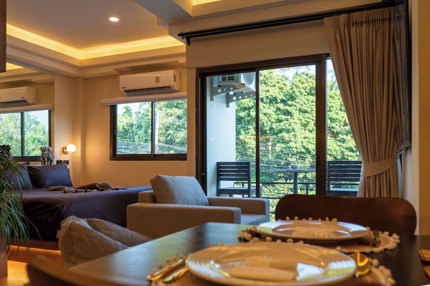 A hotel room with a bed, sofa, dining table with plates, and a balcony with a view of trees.