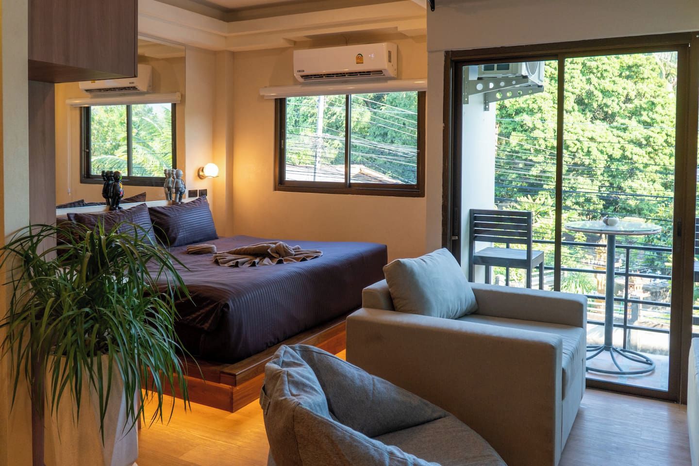 A hotel room with a bed, a sofa, a balcony with a table, and a potted plant.