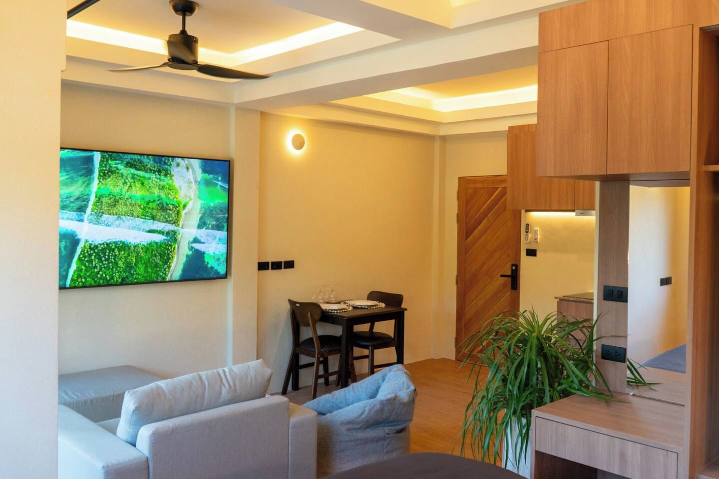 A modern living room with a flat-screen TV, a ceiling fan, a dining area with a table and chairs, and a wooden door.