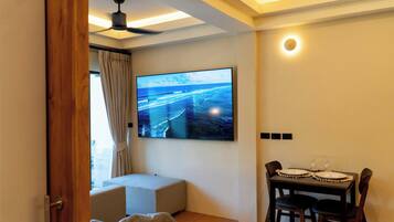A room with a flat-screen TV mounted on the wall, a dining table with chairs, a ceiling fan, and a window with curtains.