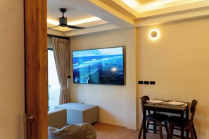 Una habitación con una televisión de pantalla plana montada en la pared, una mesa de comedor con sillas, un ventilador de techo y una ventana con cortinas.