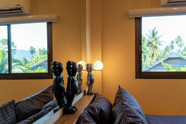 Une chambre avec un lit, une fenêtre donnant sur des palmiers et une lampe fixée au mur.