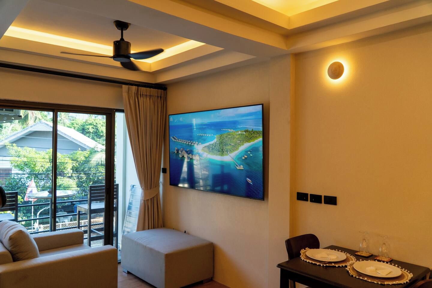 A hotel room with a large window, a ceiling fan, a flat-screen TV, a dining table with plates and glasses, and a cushioned seating area.