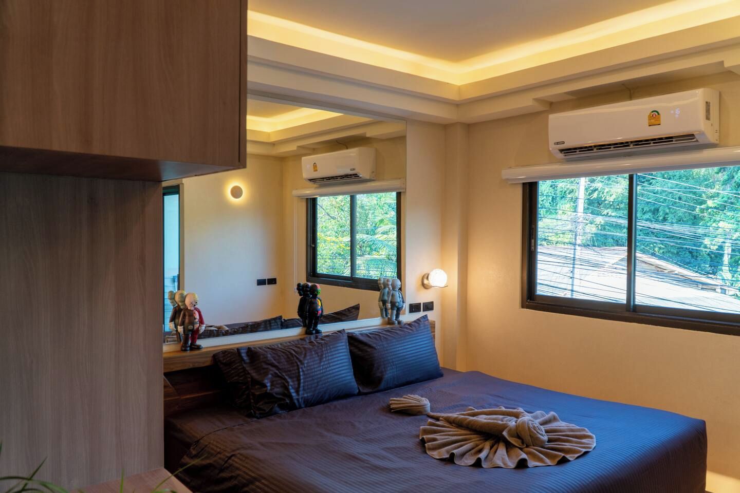 A modern bedroom with a large bed, a window with a view, an air conditioning unit, and a built-in bench with decorative items.