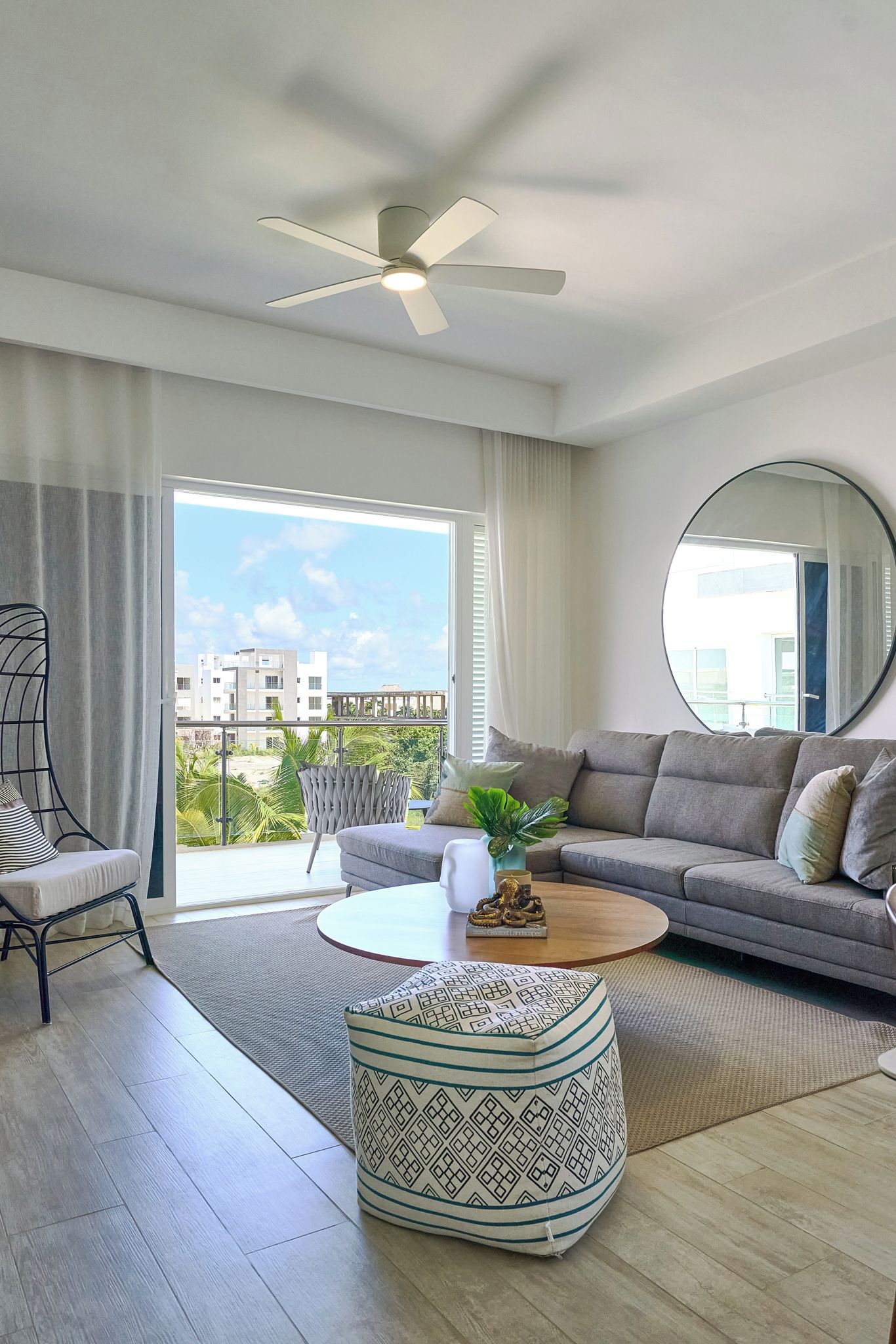 Ein modernes Wohnzimmer mit einem grauen Sofa, einem runden Couchtisch, einem gemusterten Hocker, einem Deckenventilator und einem großen Fenster mit Blick auf Gebäude und Grünflächen.