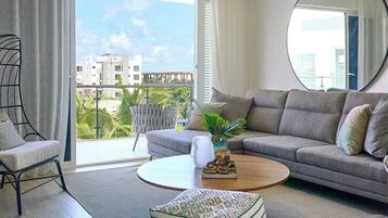 Ein modernes Wohnzimmer mit einem grauen Sofa, einem runden Couchtisch, einem gemusterten Hocker, einem Deckenventilator und einem großen Fenster mit Blick auf Gebäude und Grünflächen.