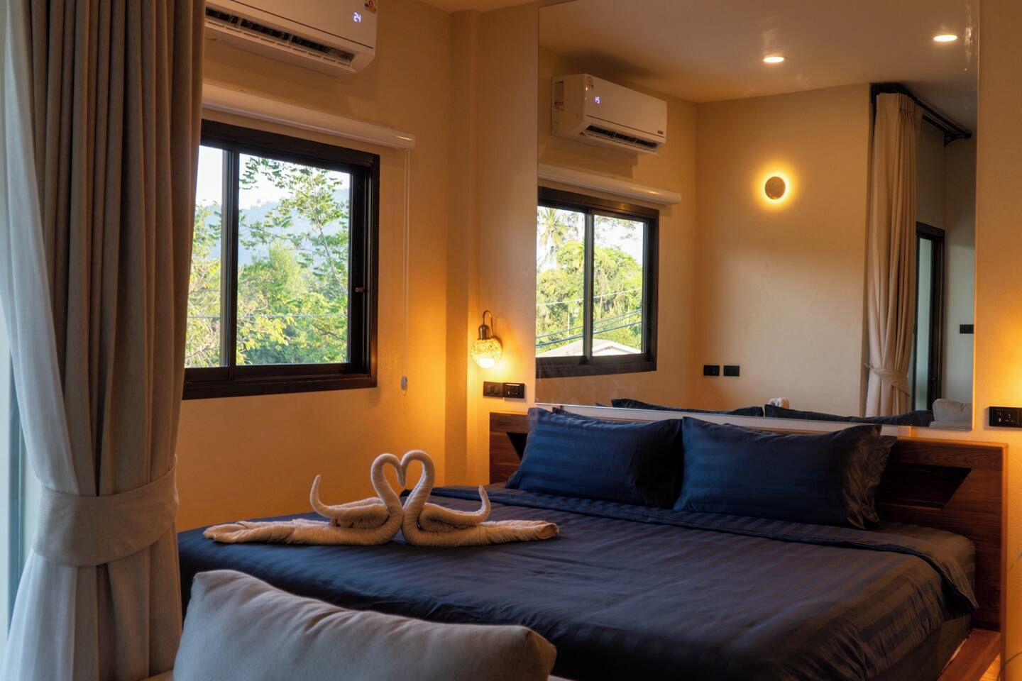 A hotel room with a bed, two windows, an air conditioning unit, and a wall-mounted lamp.