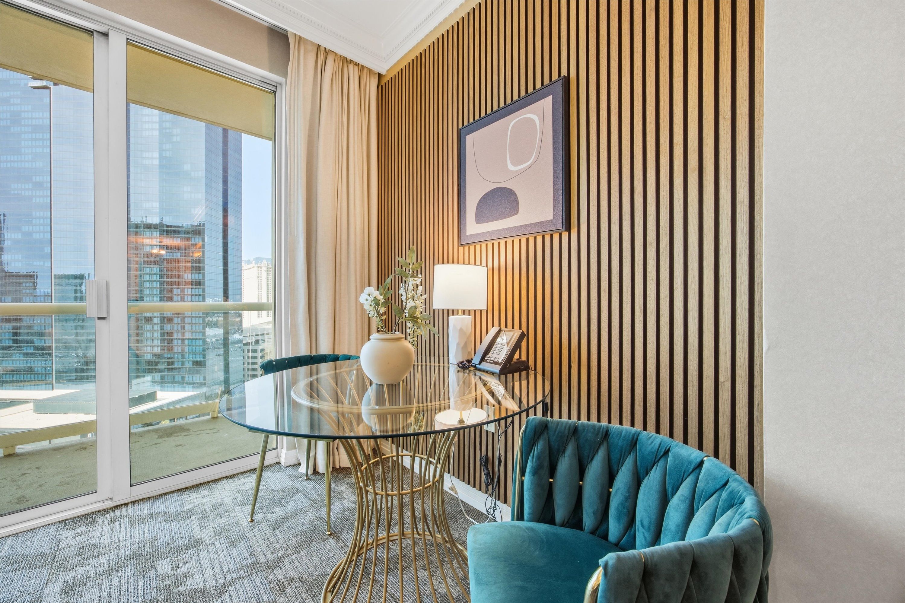 Ein modernes Hotelzimmer mit Glastisch, blauem Sessel, einer Wand aus Holzlamellen und Blick auf die Stadt.