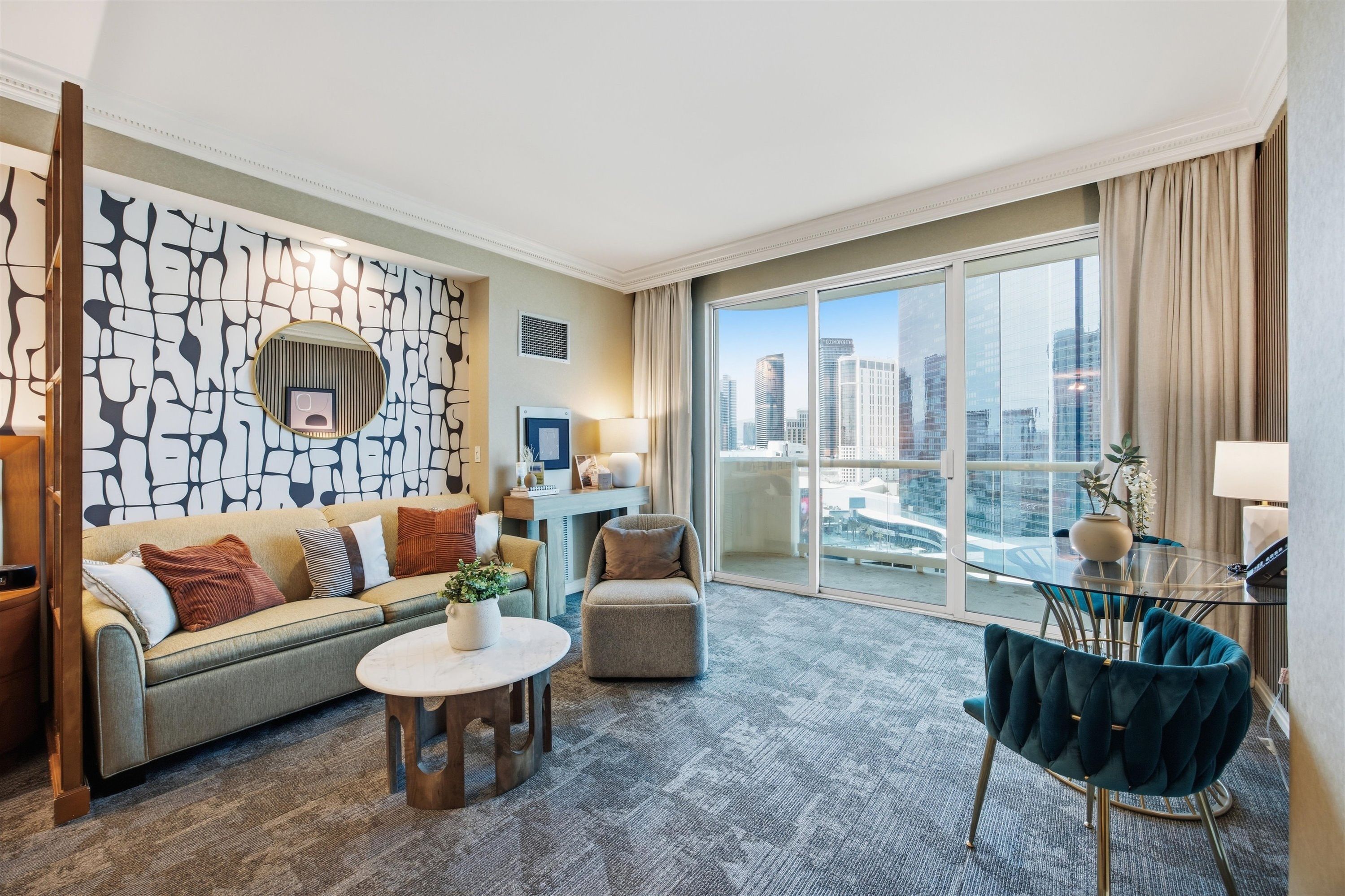 Una habitación de hotel moderna con un gran ventanal que ofrece vistas a la ciudad, un sofá con cojines decorativos, una mesa de centro redonda y un sillón tapizado en azul.