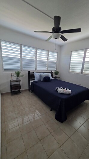 A bedroom with a bed, a ceiling fan, a nightstand with a plant, and a window with blinds. - BRISAS - Lovely 3-bedroom house with AC in cool Boquerón (BOQUERÓN)