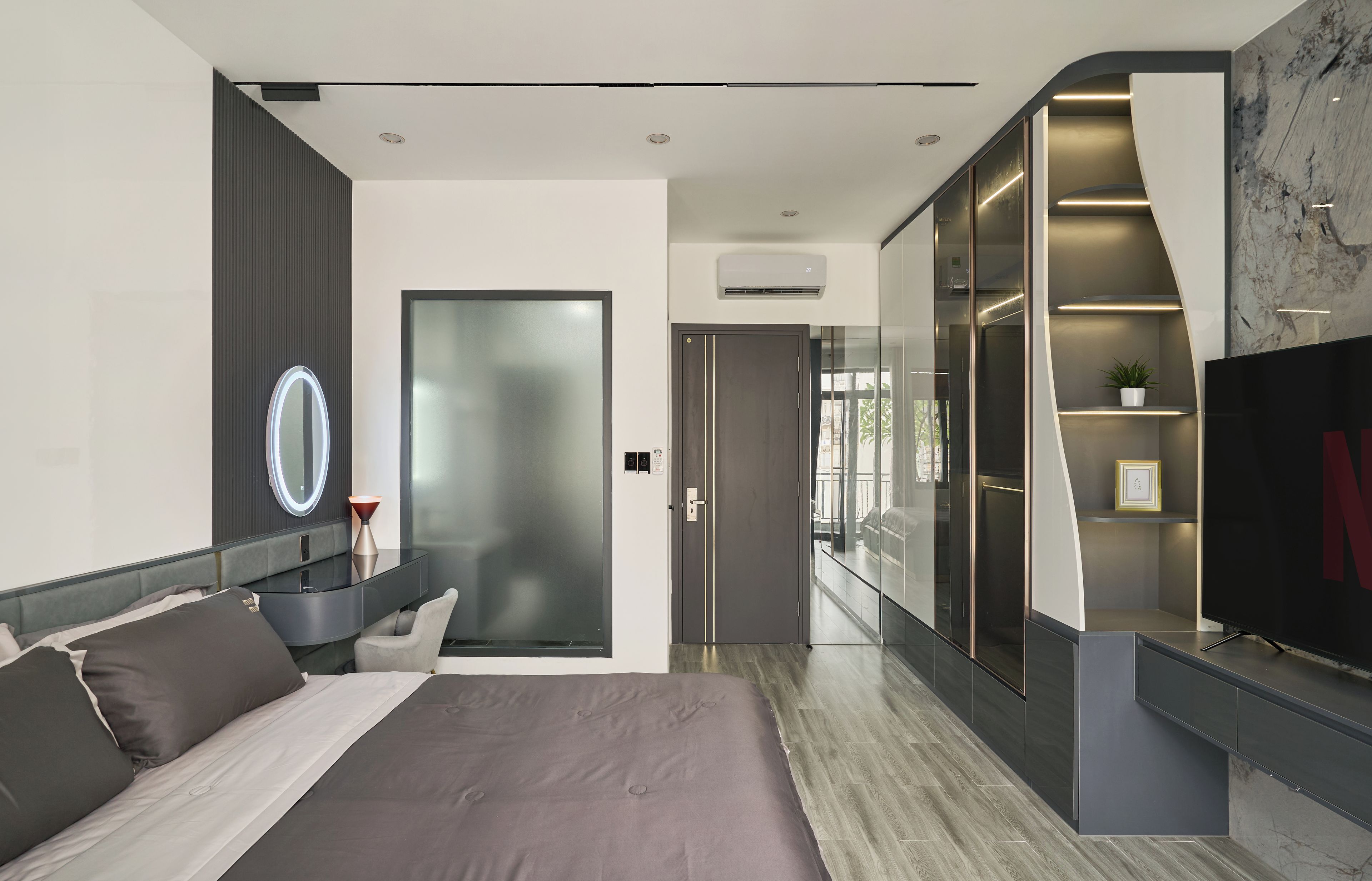 A modern bedroom with a bed, bedside table, mirror, and a wardrobe with a built-in TV.