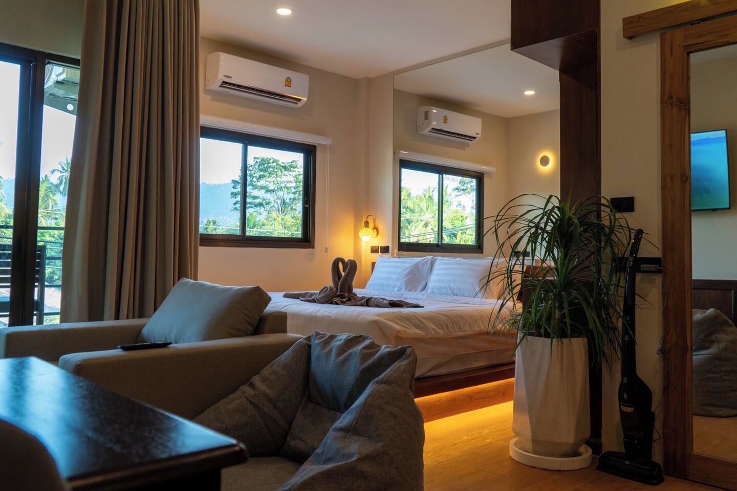 A hotel room with a bed, a sofa, a potted plant, and two air conditioning units.