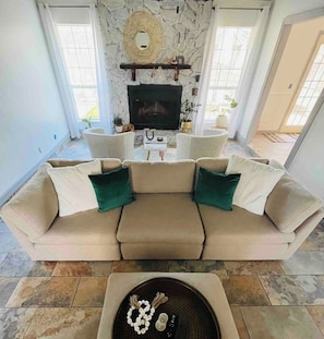 A living room with a beige sectional sofa, a fireplace, and a stone wall. - Charming 3-bedroom house with pool and spacious backyard (Lynn Haven)