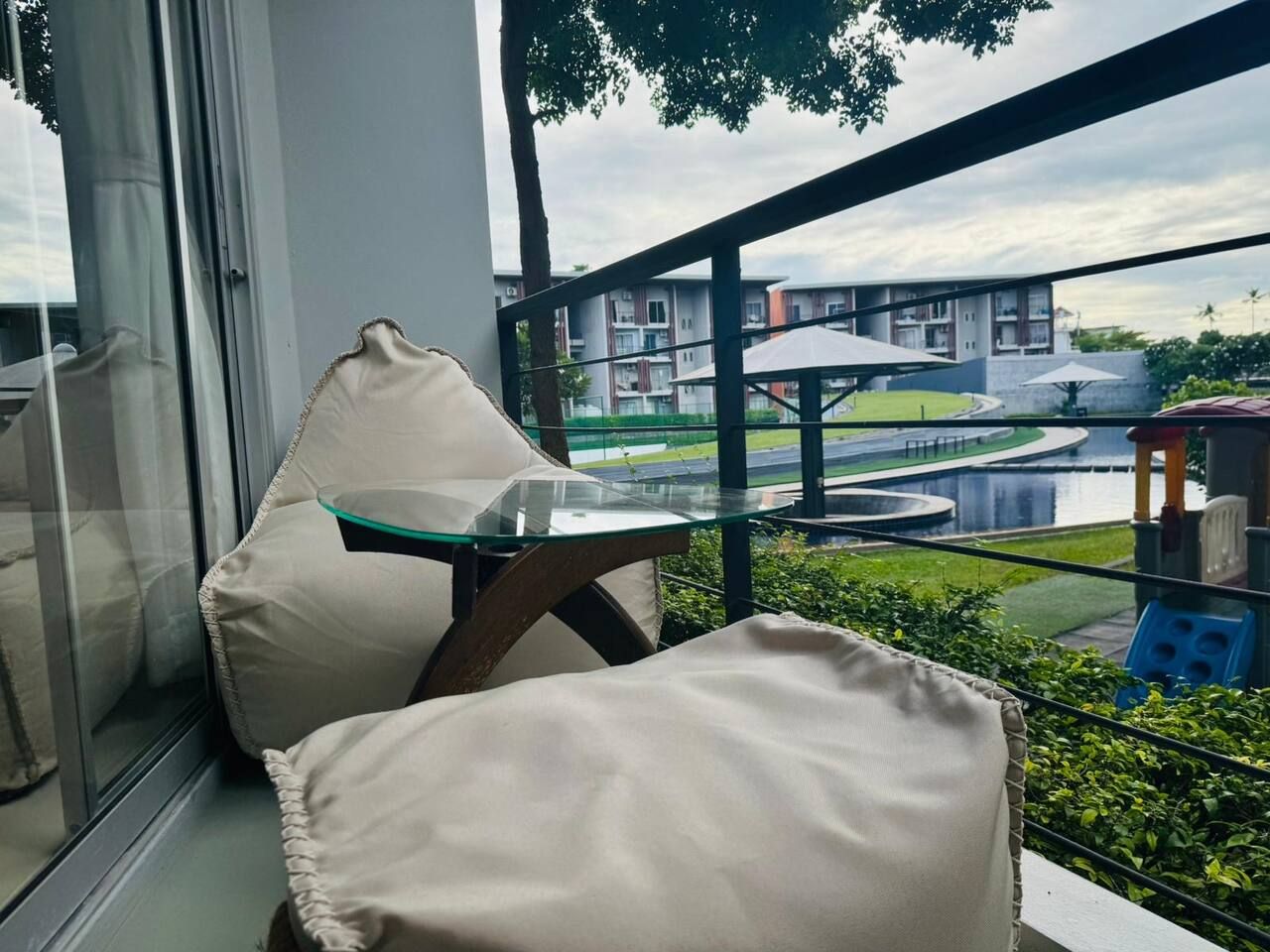 A balcony with a glass table and chairs, overlooking a landscaped area with a building in the background.