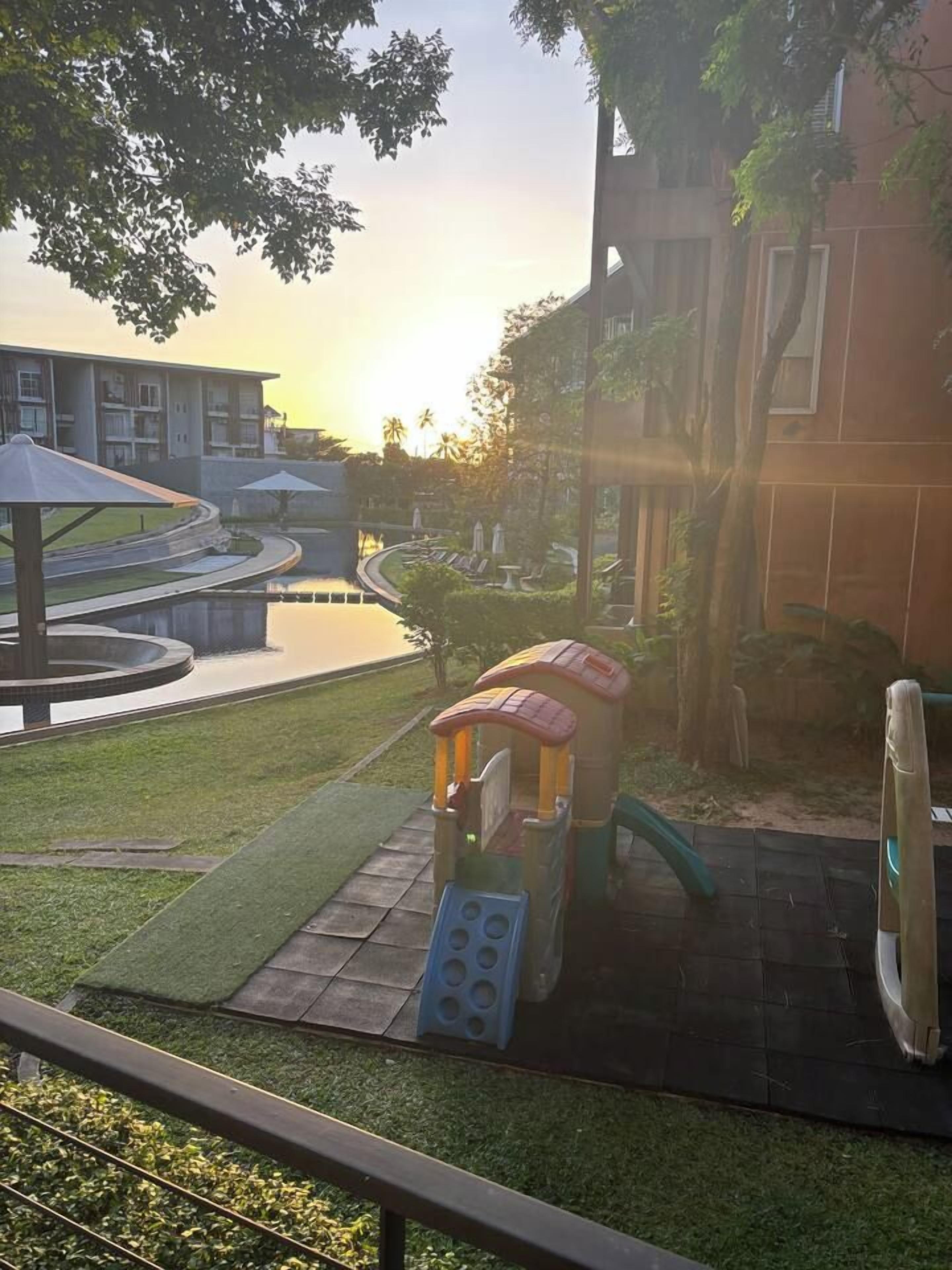 Parque infantil com escorrega e estrutura de escalada, rodeado por área ajardinada com fonte de água e um edifício ao fundo.