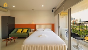 Quarto de hotel com cama, sofá verde, pequena mesa com frutas, máquina de lavar e varanda com vista para a cidade.