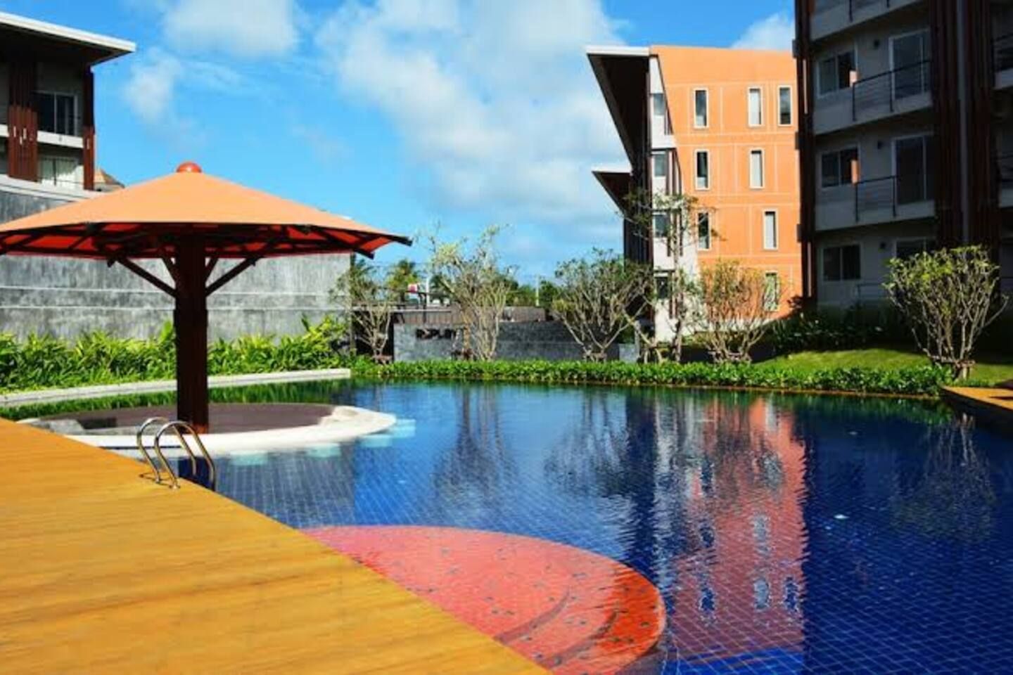 Une piscine dotée d’une terrasse en bois et d’un motif circulaire rouge, entourée de verdure et de bâtiments modernes.