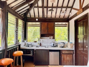 A compact kitchen with a sink, stove, and refrigerator, wooden cabinets, and a window with a view of greenery.