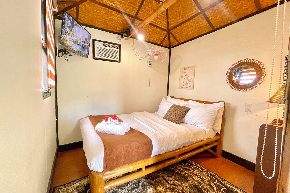 A single bed with a wooden frame, a wall-mounted air conditioner, a ceiling fan, a woven ceiling, and a patterned rug.