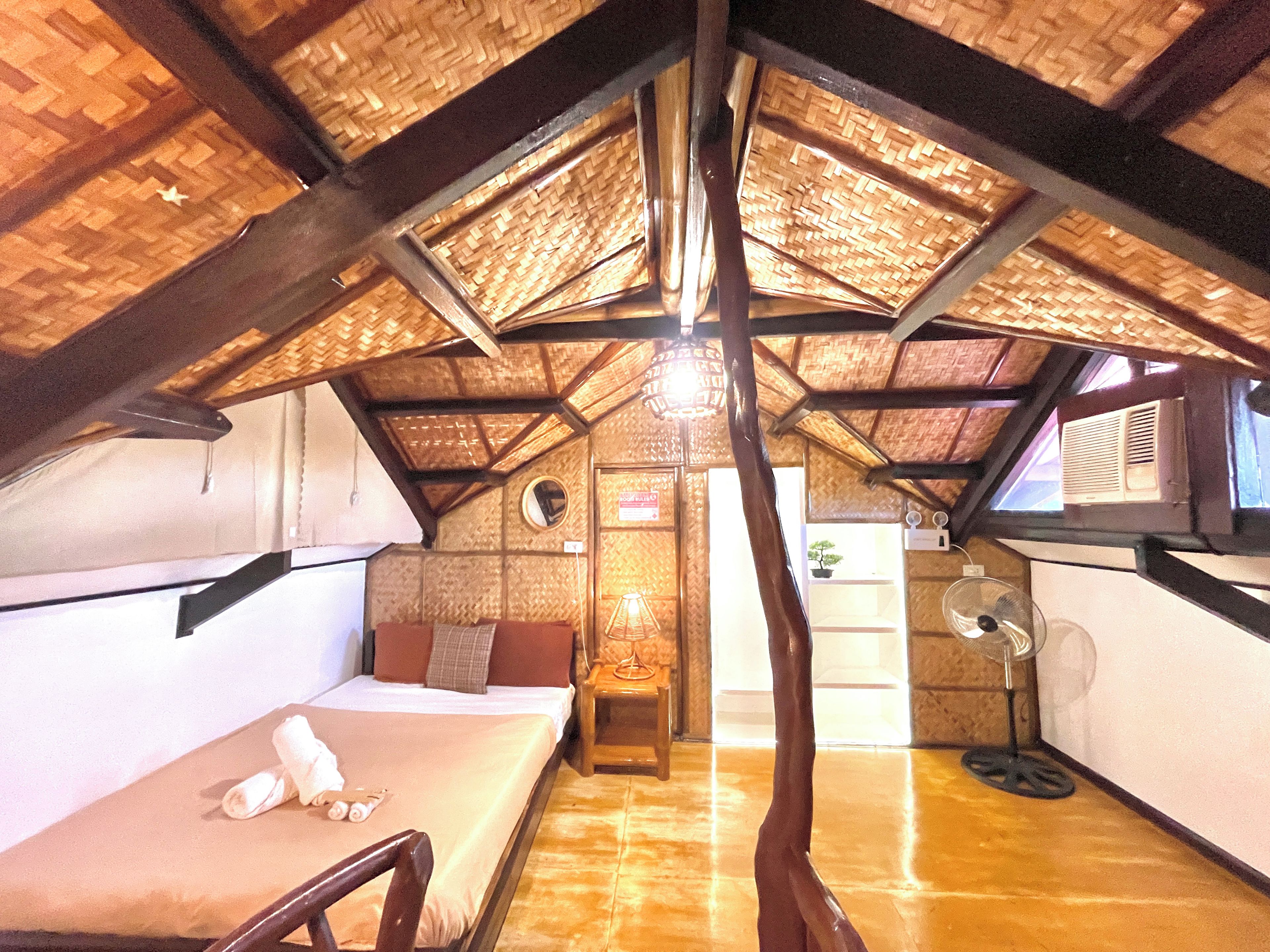 A room with a sloped ceiling, a bed, a chair, a table, a fan, and a window.