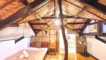 A room with a sloped ceiling, a bed, a chair, a table, a fan, and a window.