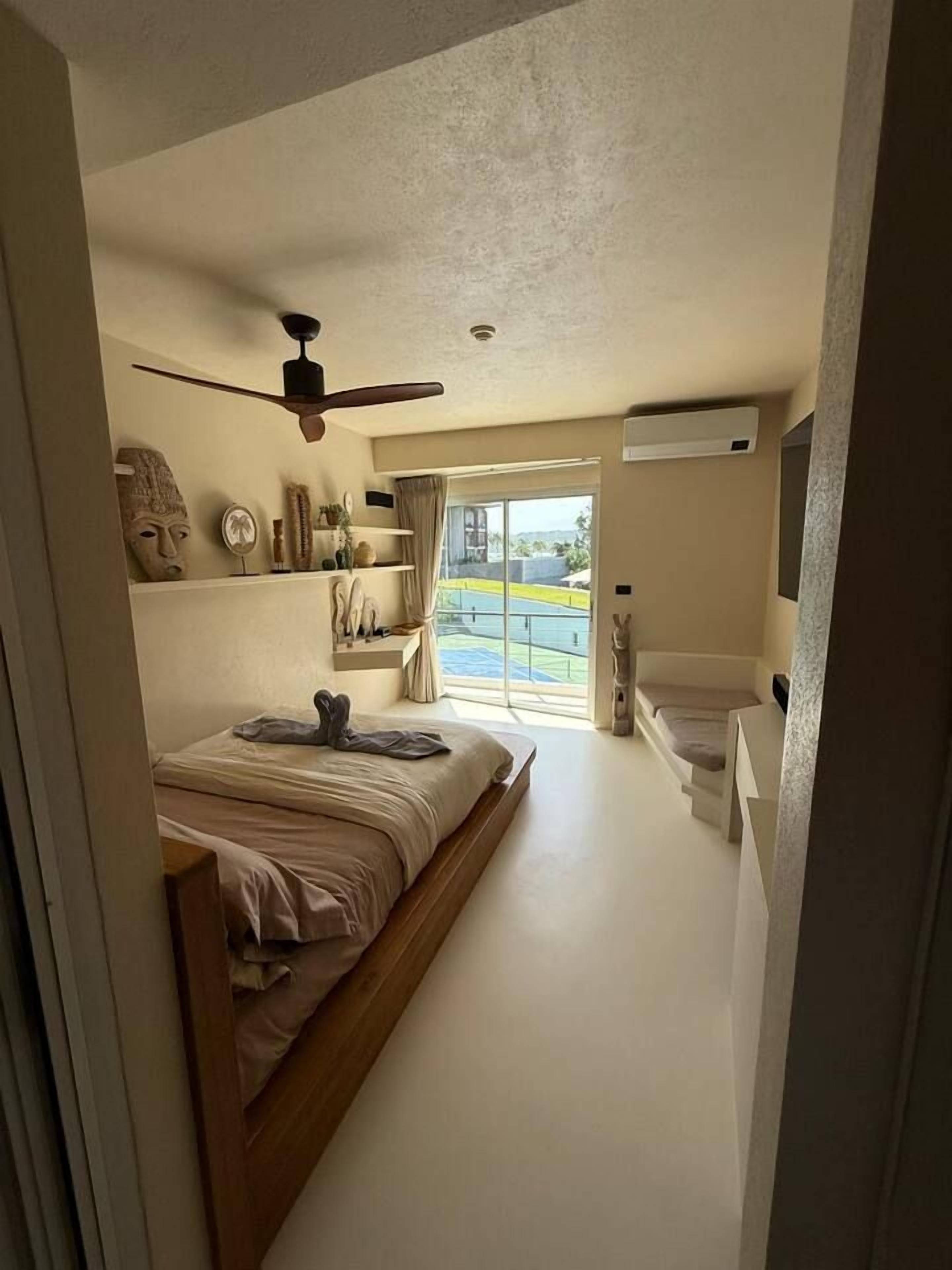 Une chambre à coucher avec un lit, un ventilateur de plafond, une fenêtre avec des rideaux et une vue sur l’extérieur.