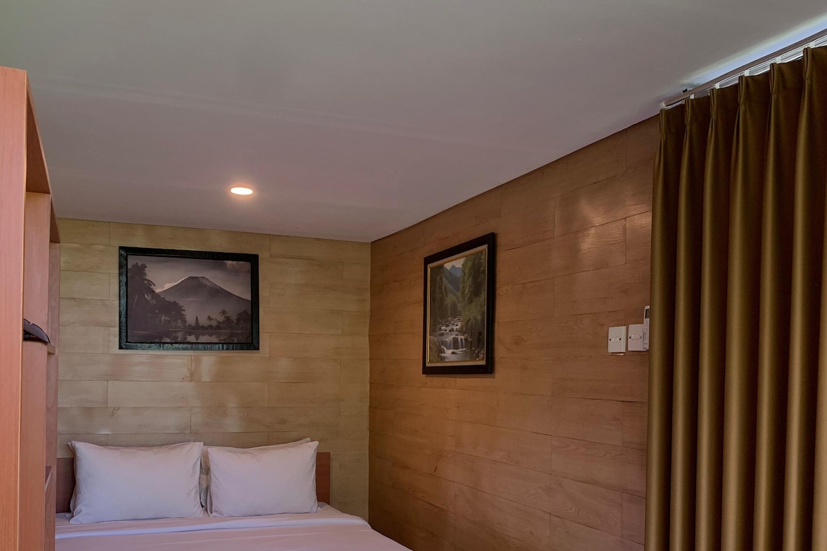 A bedroom with a wooden bed, two pillows, a headboard, a wooden floor, a wall with two framed pictures, and a curtain.