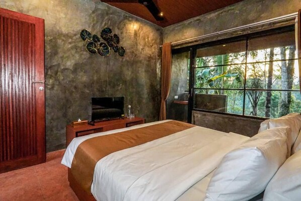 A bedroom with a bed, a television, a window with a view of trees, and a wooden door.