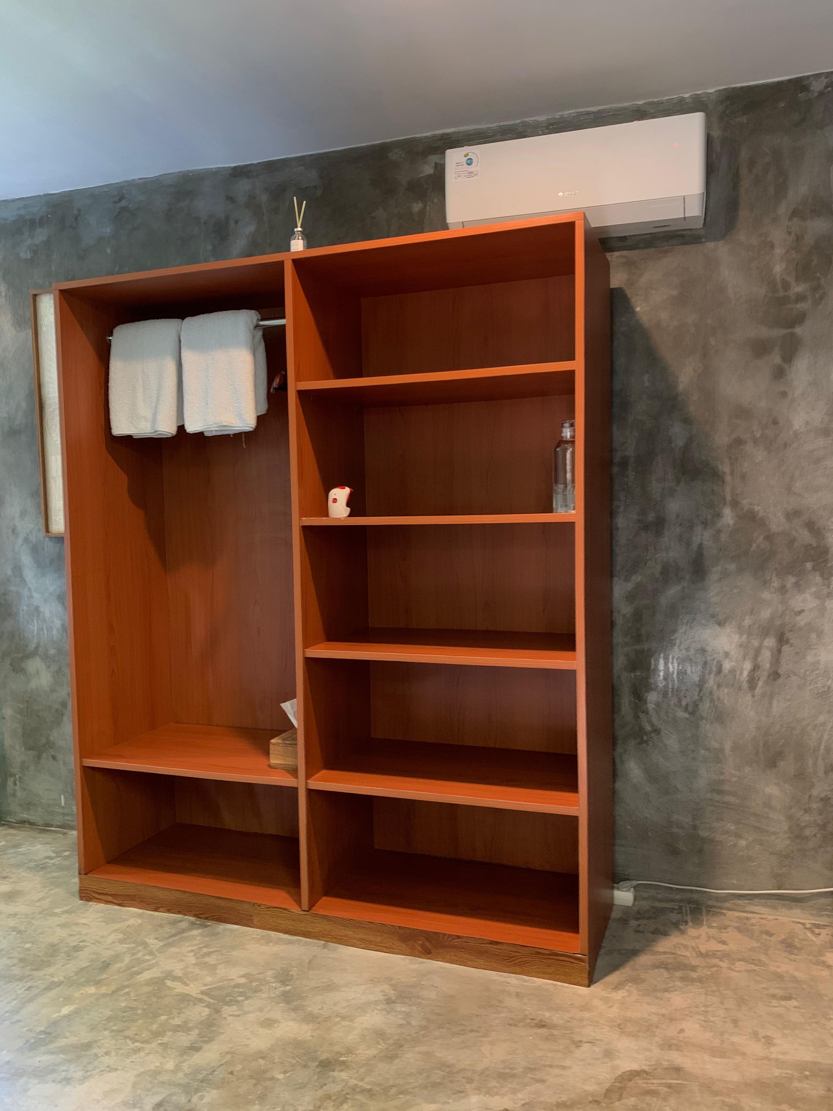 A wooden wardrobe with multiple shelves and hanging space, a wall-mounted air conditioner, and a concrete floor.