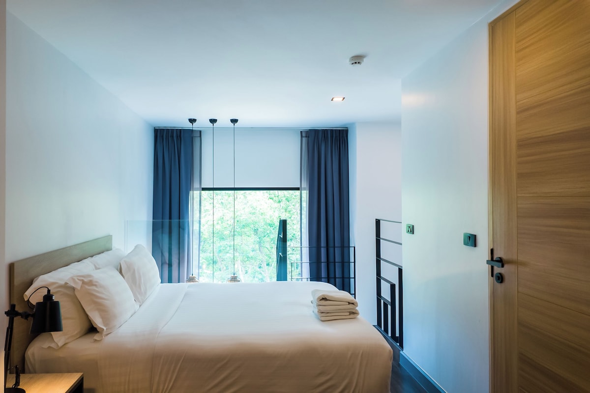 A hotel room with a bed, a nightstand, a window with curtains, a door, and a ceiling light.