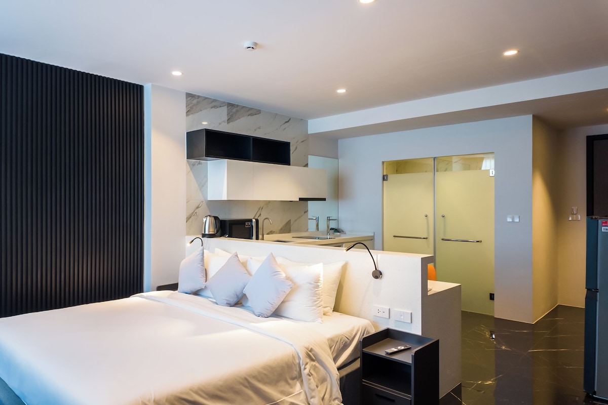 A modern hotel room with a large bed, a built-in desk, a kitchen area, and a bathroom with a mirror.