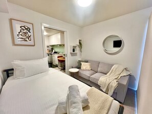 A compact room with a bed, a sofa, a small table, and a round mirror. There is a kitchenette visible through an open door. - Modern Box Hill Studio w Balcony & Great Location (Box Hill)