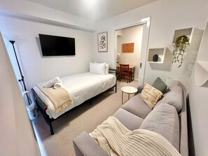A modern, well-lit room with a bed, a sofa, a small table, and a TV mounted on the wall. - Modern Box Hill Studio w Balcony & Great Location (Box Hill)