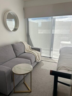 A modern living room with a grey sofa, a round coffee table, a mirror, and a view of the city through large windows. - Modern Box Hill Studio w Balcony & Great Location (Box Hill)