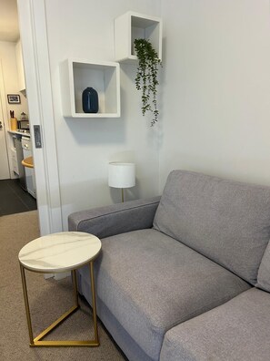 A modern living room with a grey sofa, a small round table with a gold frame, a white floor lamp, and a white shelf with a hanging plant. - Modern Box Hill Studio w Balcony & Great Location (Box Hill)