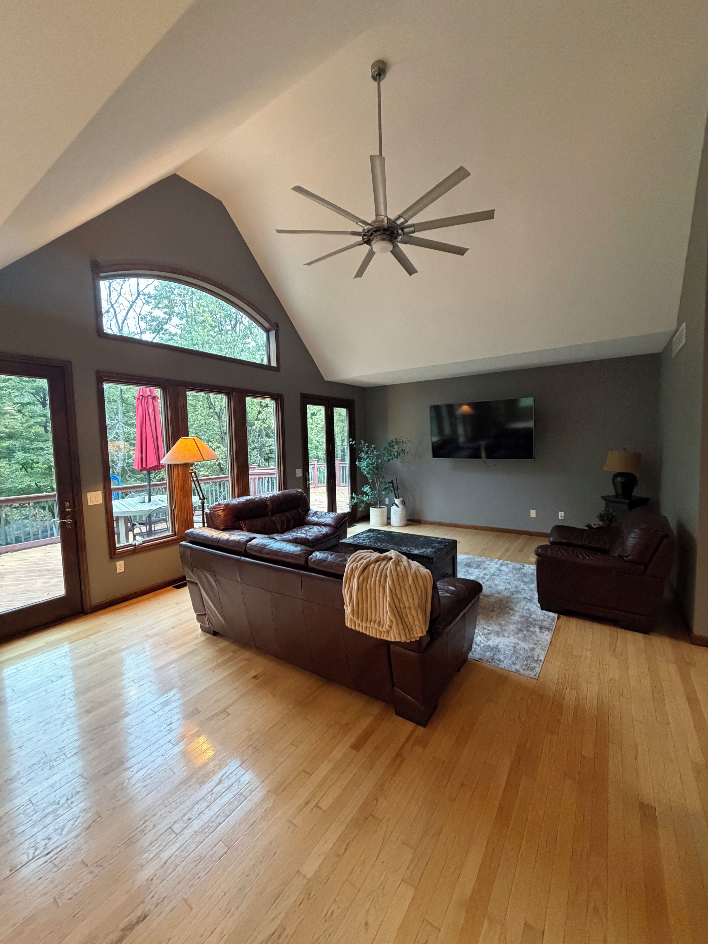 A spacious living room with a large brown leather sofa, a ceiling fan, a flat-screen TV, and a view of a deck through large windows.