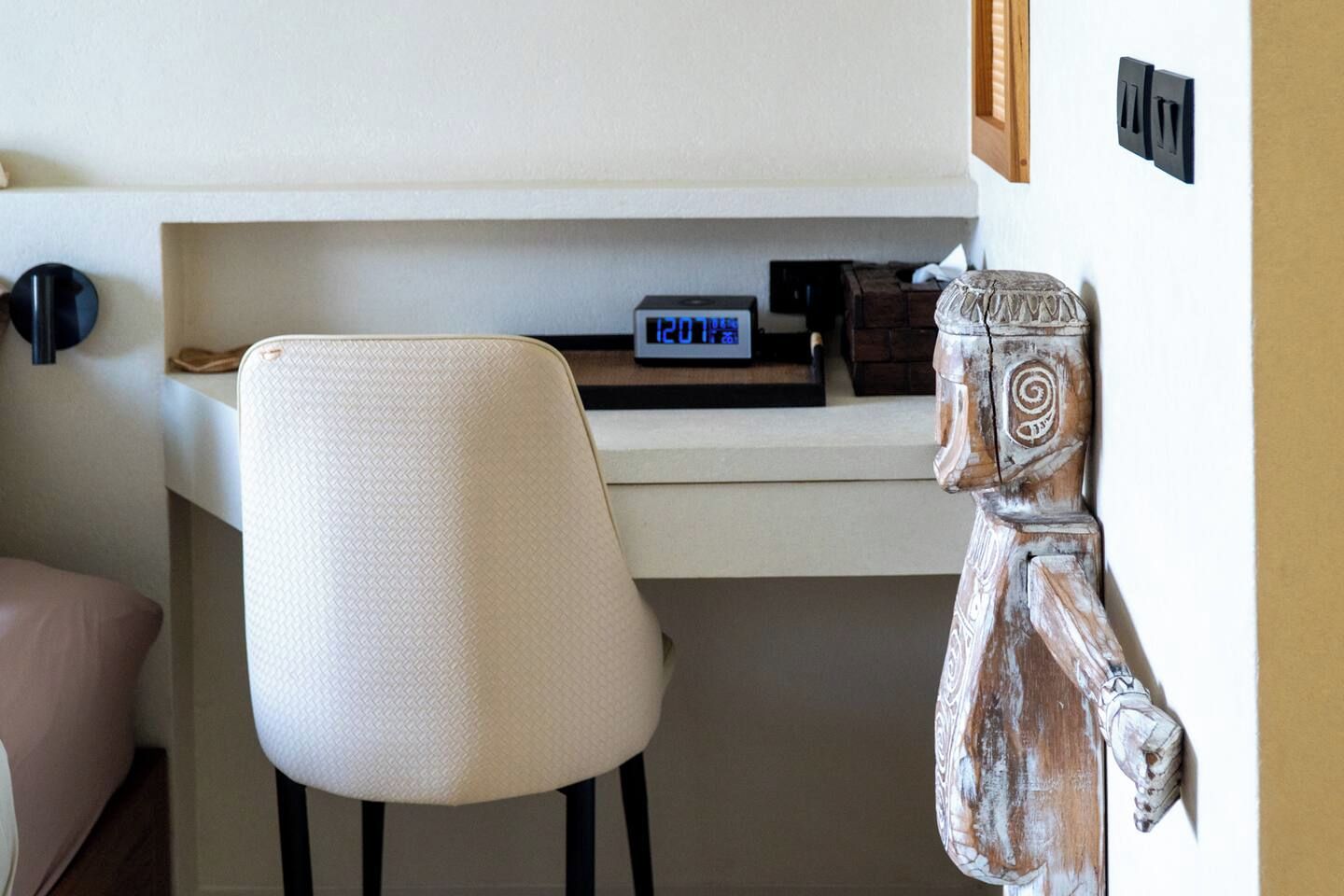 A room with a white chair, a wooden sculpture, and a digital clock on a shelf.