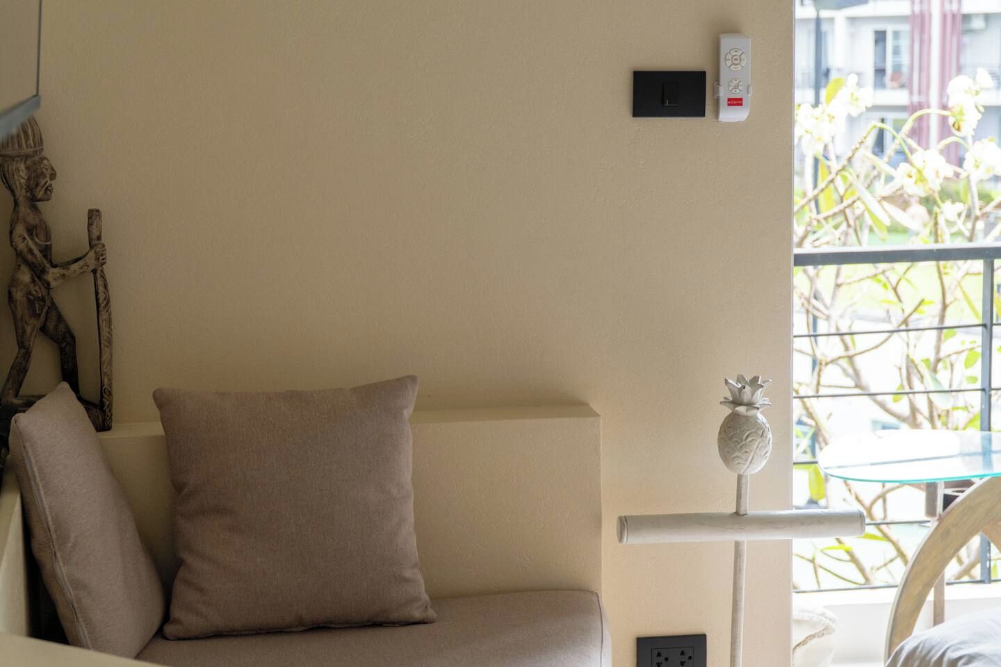 A room with a beige sofa, a small table with a decorative item, a wall-mounted thermostat, and a window with a view of trees.