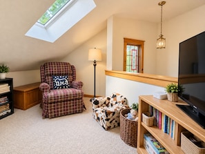 A cozy room with a high ceiling, a skylight, a plaid armchair, a cow-print ottoman, a bookshelf, and a television. - 4BR Poconos Retreat, Fall at Camelback (Tannersville)