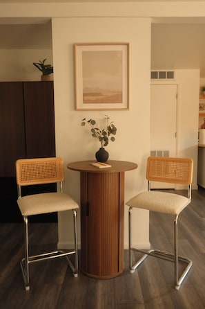 Un comedor con dos sillas, una mesa redonda, un florero con planta y un cuadro en la pared.