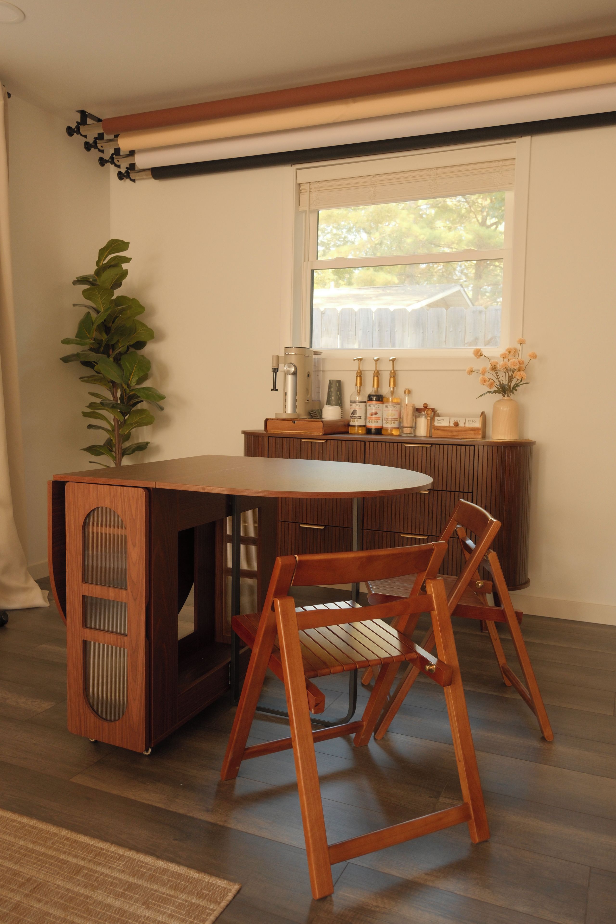 A dining area with a wooden table and chairs, a potted plant, and a window with a view of trees.