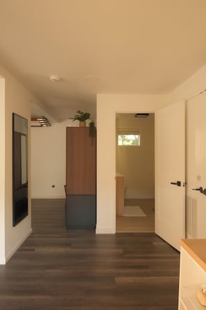 Un pasillo con suelo de madera, una puerta que da a una habitación con ventana, y una planta sobre un mueble.