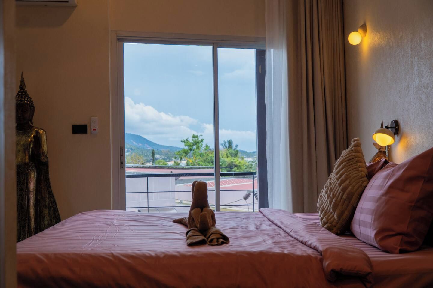 A hotel room with a bed, a statue, a view through a large window, and a wall-mounted lamp.