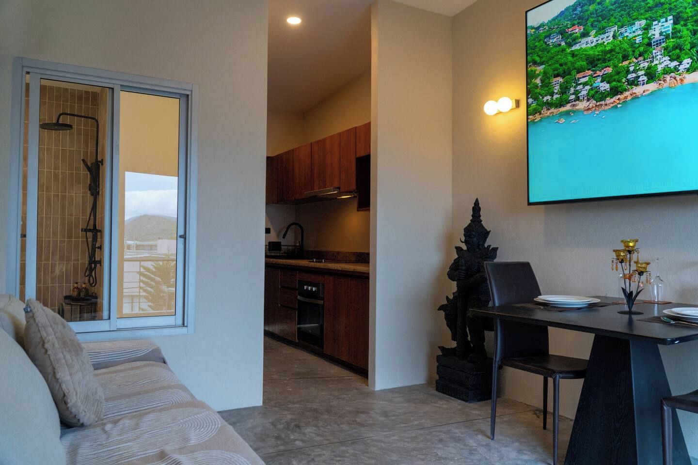 A modern living room with a sofa, a dining table with chairs, a kitchen in the background, and a large TV screen displaying a scenic view.