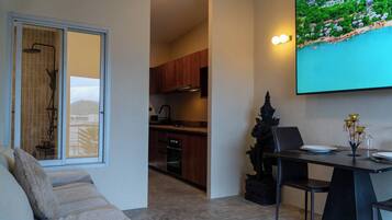 A modern living room with a sofa, a dining table with chairs, a kitchen in the background, and a large TV screen displaying a scenic view.
