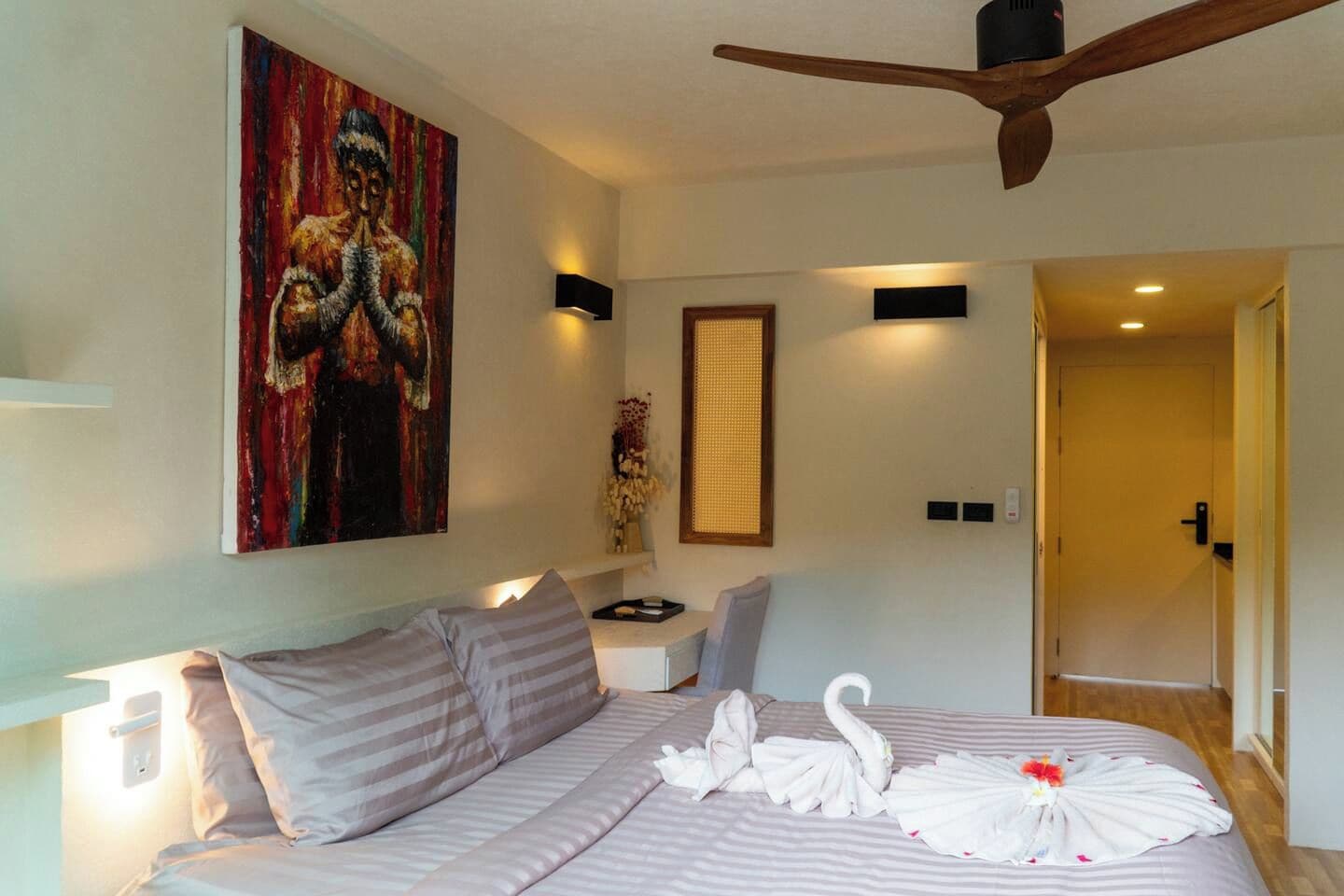 Une chambre d’hôtel avec un lit, un tableau, un ventilateur de plafond et une porte donnant sur une autre pièce.