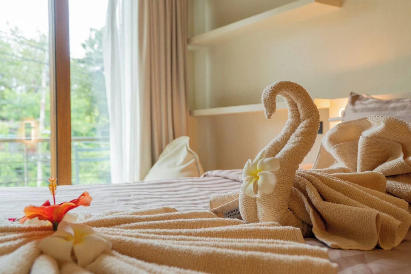 A hotel room with a bed, a window with curtains, and a towel with a flower.