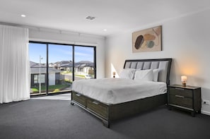 A bedroom with a large bed, a bedside table with a lamp, and a view of a residential area through a sliding door. - Family 5-bed House Near Tarneit Central (Melbourne)