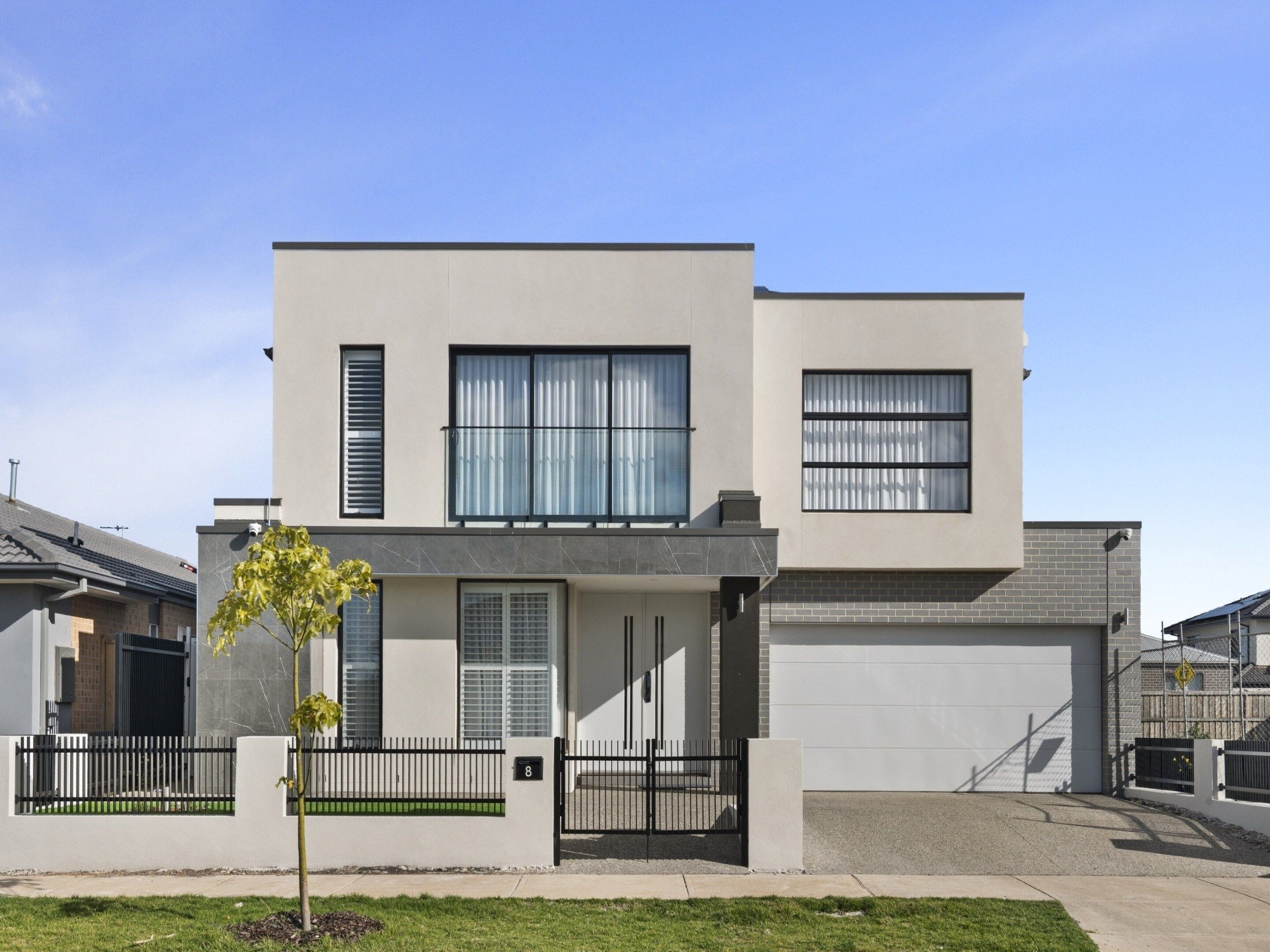 Une maison moderne à deux étages, dotée d’un garage, d’un petit jardin devant et d’un arbre.